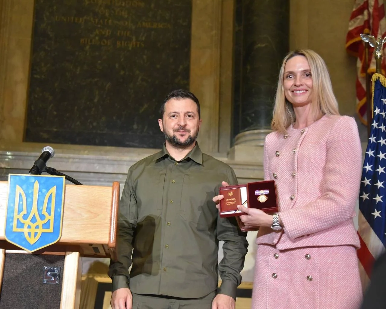 Cerimonia di premiazione: una donna in giacca rosa tiene un riconoscimento accanto a un uomo in camicia militare